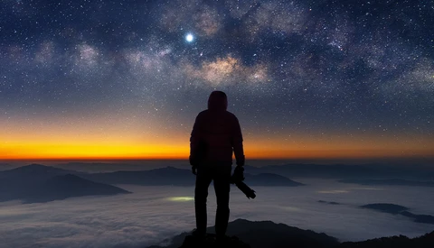 Silhouetted figure standing on a mountain peak gazing at the Milky Way and sunrise.