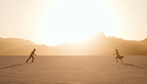 Two silhouetted figures in crouched positions on a vast sandy plain, backlit by a golden sunset behind distant mountains.
