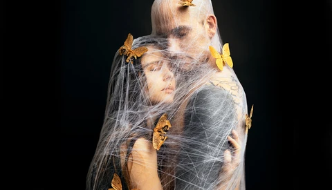 Two figures wrapped in delicate spider web with golden butterflies perched on their bodies and web.