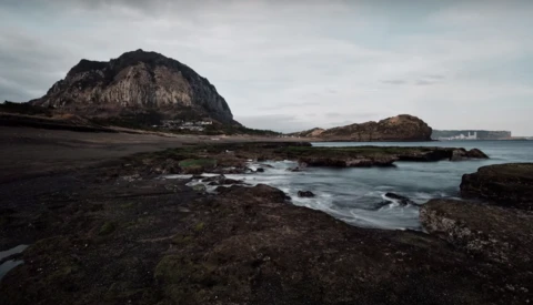 Rocky volcanic coastline with dark sand beach, prominent mountain peak, and calm ocean waters under overcast sky.