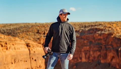 Man in cap and dark jacket holding camera stands at cliff edge overlooking layered canyon terrain.