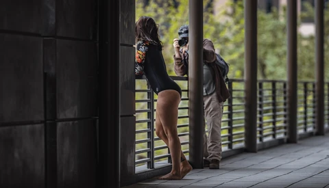 Woman standing on a wooden deck viewed through dark doorframe, with two people visible in background near railing.