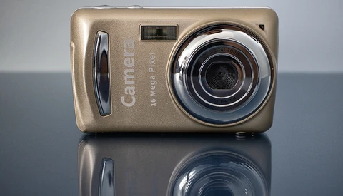 Gold compact digital camera with chrome lens bezel and grip, reflected on mirror surface against gray background.