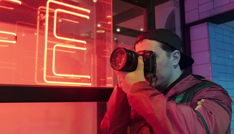 Photographer holding a digital camera with a large red lens, shooting against neon signage in a vibrant red and purple lit environment.