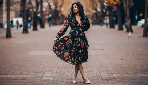Woman in black floral dress standing on a brick plaza lined with trees.