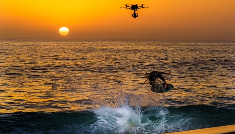 Surfer riding a wave at sunset with a drone flying overhead.