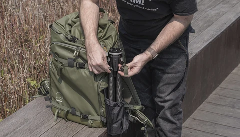 Person adjusting straps on an olive green camera backpack while standing on a wooden deck.