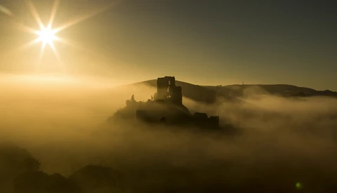 Medieval castle tower emerging from thick fog at sunrise with golden sunlight streaming overhead.