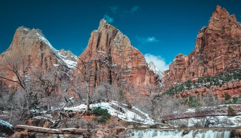 Red rock canyon with snow-capped peaks under clear blue sky and flowing water in foreground.