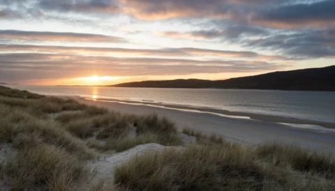 Sunset over a coastal bay with dunes in foreground and hills across the water.