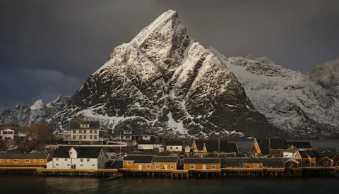 Snow-covered mountain peak rising behind a waterfront village with yellow and orange wooden buildings.