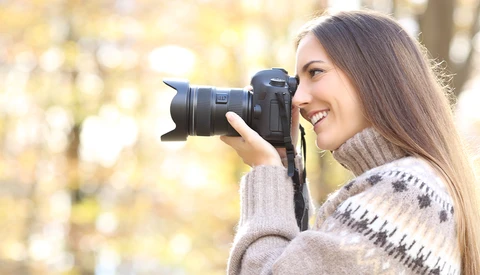 Woman holding a DSLR camera with telephoto lens, smiling while photographing outdoors.