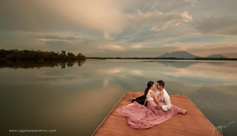 Couple sitting together on a wooden dock overlooking a calm lake with mountains and sky reflected in the water.
