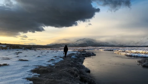 Solitary figure standing on rocky shore at golden hour with snow-capped mountains and dramatic cloudy sky in distance.