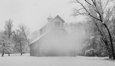 Rural stone barn surrounded by bare trees during heavy snowfall with low visibility.
