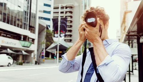 Man in light blue shirt and tie holding VR headset to his face on an urban city street.