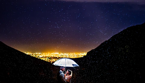Couple holding illuminated umbrella in rain beneath starry night sky with city lights below.