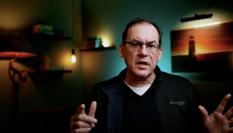Man wearing glasses and dark jacket gestures while speaking in a studio setting with warm ambient lighting.