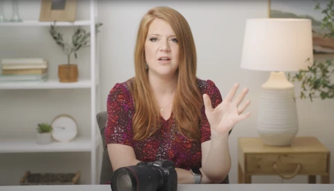 Woman with reddish-brown hair in burgundy patterned shirt gesturing while speaking in home interior setting.