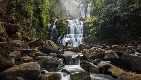 Multi-tiered waterfall cascading over moss-covered rocks in a lush forest canyon with towering vegetation.