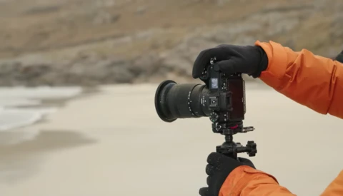 Photographer in orange jacket holding DSLR camera on tripod at beach during overcast conditions.