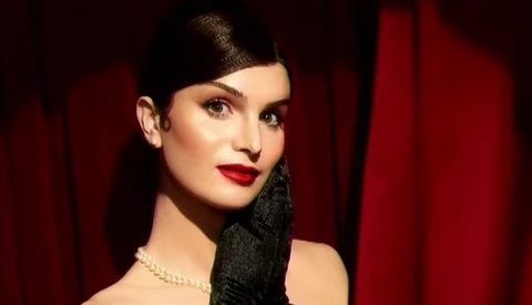 Woman in profile wearing red lipstick and black gloves against dark red curtain backdrop.