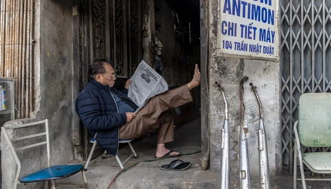 Man reading newspaper while sitting in doorway of weathered storefront with metal security gate.