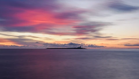 Seascape at sunset with dramatic pink and purple clouds reflecting on calm water and a distant lighthouse on the horizon.