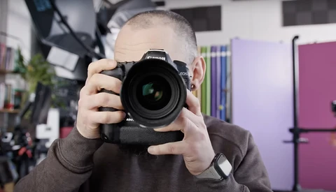 Young photographer holding a DSLR camera with battery grip up to his face in a studio setting.