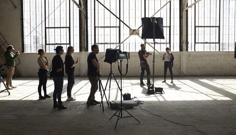 Film crew setting up camera and lighting equipment in an industrial studio space with large windows.