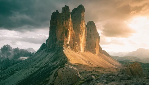 Dramatic mountain peaks with vertical columnar formations lit by golden sunlight against stormy clouds.