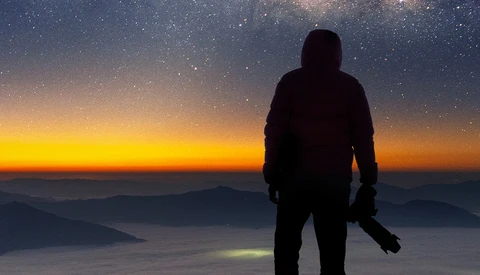 Silhouetted figure standing on a mountain ridge at sunrise, gazing toward a starry sky and golden horizon above layered clouds.
