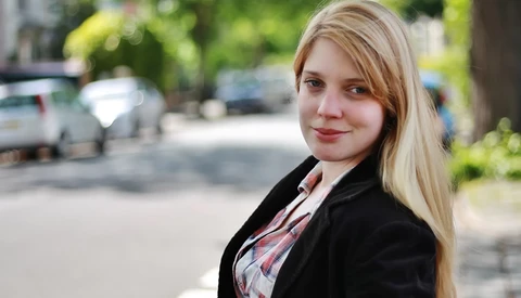 Woman with blonde hair wearing a black blazer and plaid shirt, smiling at camera outdoors.