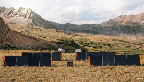 Solar panel array set up in a mountain valley with dramatic peaks and green meadows in background.