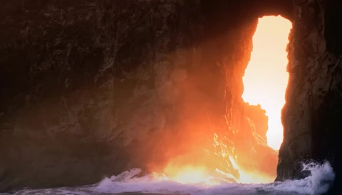 Brilliant sunlight streams through a narrow cave opening onto crashing waves and rocky shores.