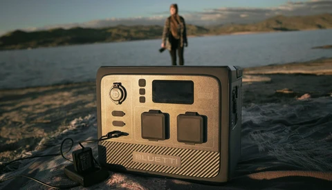 Portable power station with tan and black casing displayed on a beach, with a person standing in shallow water in the background.