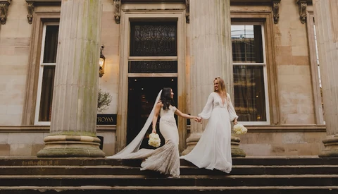 Two brides in flowing white wedding gowns with dramatic trains pose on classical stone steps holding bouquets.