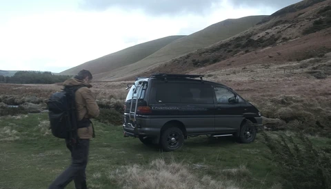 Hiker with backpack standing beside a black van parked on moorland with rolling hills in background.