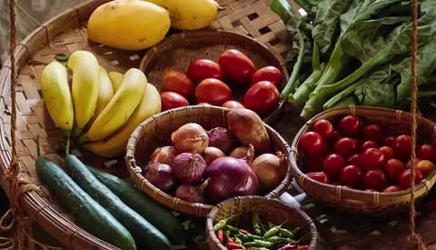 Overhead view of fresh produce arranged in woven baskets, including bananas, potatoes, tomatoes, onions, cucumbers, and green peppers.