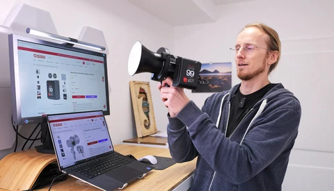 Photographer holding a cinema camera while reviewing footage on dual monitors at a wooden desk.