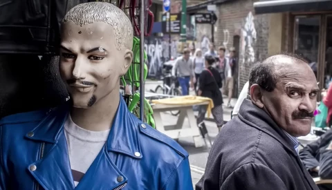 Man with mustache looking over shoulder at camera, street scene with mannequin head in blue jacket visible in background.