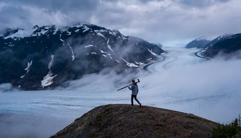 Hiker standing on a rocky outcrop overlooking a vast glacier valley surrounded by snow-capped mountains.