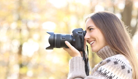 Woman holding a DSLR camera with telephoto lens, photographing outdoors with warm backlighting.