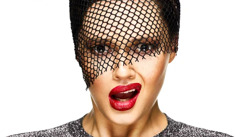 Portrait of a person wearing a black mesh veil covering the eyes, with bold red lipstick and a sparkly dark top against a white background.