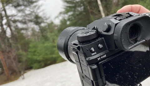 Fujifilm X-H2 mirrorless camera held in hand outdoors with forest backdrop.