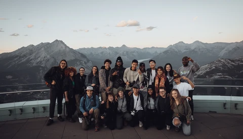 Large group of people posing together on an elevated platform with snow-capped mountains in the background during golden hour.