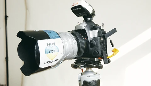 Professional video camera mounted on tripod with white tape reading "PRAY FOR UKRAINE" wrapped around lens barrel.