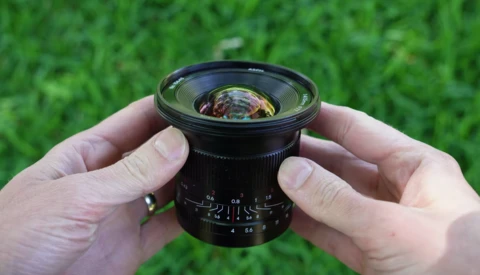Hands holding a wide-angle lens with visible aperture scale and focus ring against a green grass background.