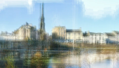 Medieval cathedral with tall spire reflected in calm river water, surrounded by historic buildings.