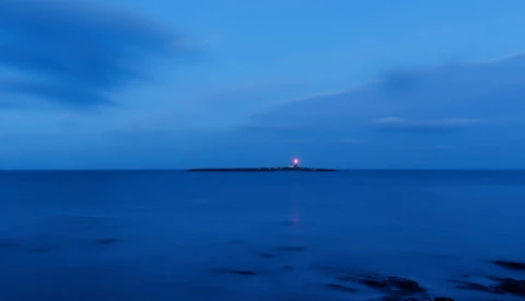 A solitary lighthouse beam cuts through deep blue twilight across a calm ocean horizon.